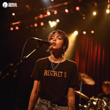 A white female recording artist is passionately singing into a microphone on stage, with a drummer playing behind her. The scene captures the intensity and emotion of her performance, emphasizing the theme of "Regret U" as she pours her heart into the music. The setting highlights the connection between the artist and her audience, conveying a sense of vulnerability and raw emotion that resonates through the atmosphere.
